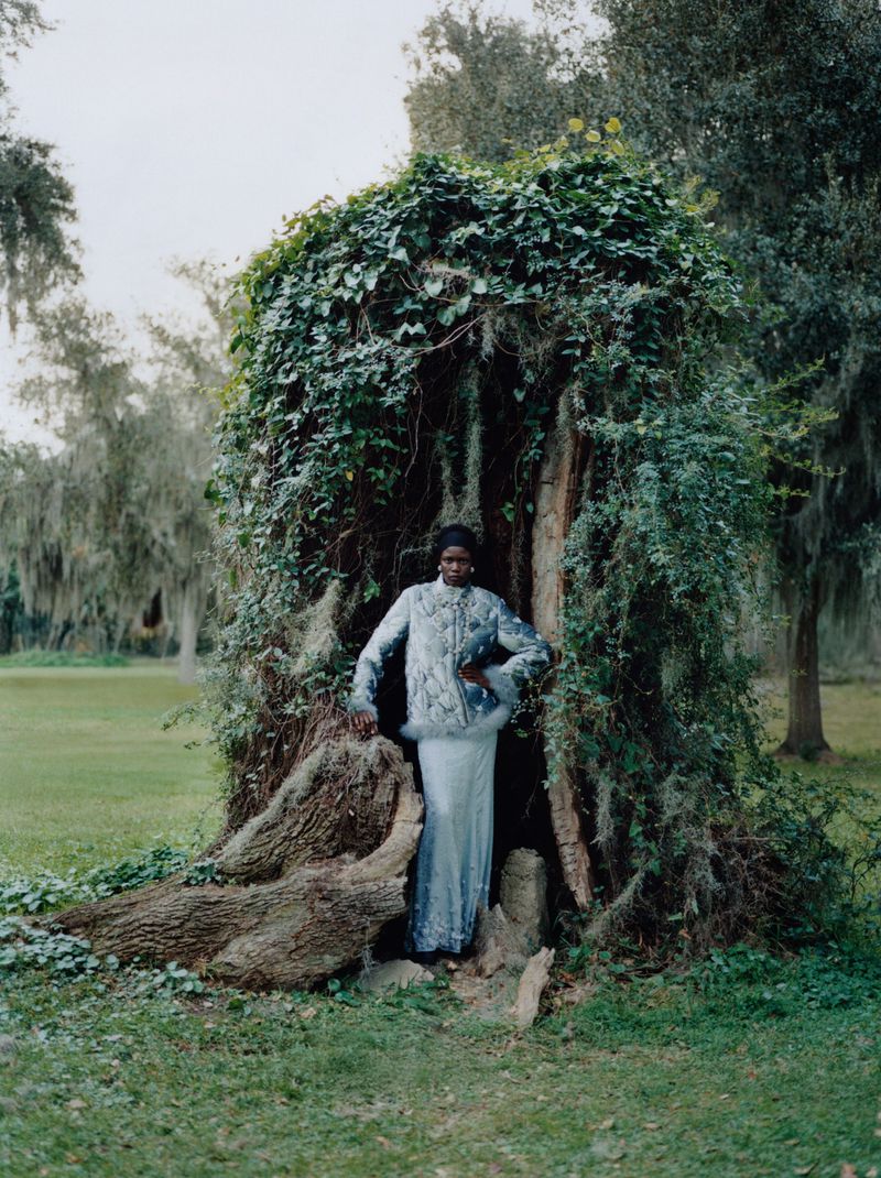 A Black woman in a blue beaded jacket and white dress poses inside a vine-covered tree trunk in a green grassy landscape.