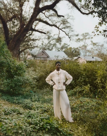 An adult female model with an afro poses in a field wearing a cream-colored blouse and cream-colored pants, standing in front of lush greenery.