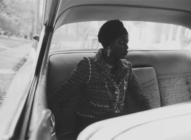 Black and white fashion shot of a woman in a car wearing a tweed jacket, earrings, and necklace, looking out the window.