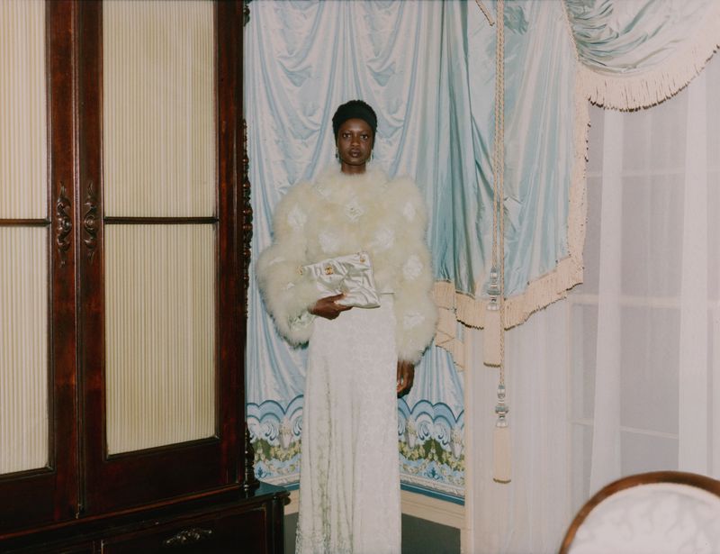 An elegant Black woman posing indoors wearing a white, feathered jacket and dress, holding a purse, set against a curtained backdrop.