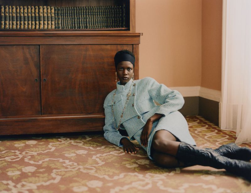 Fashion shot of an African-American woman in a tweed skirt suit and black boots, lying on a patterned rug with a wooden cabinet in the background.