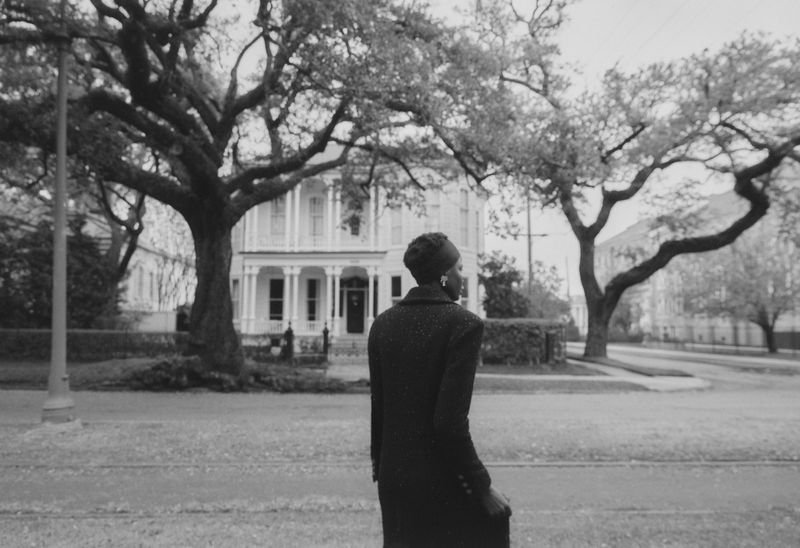 Monochrome portrait of a Black woman in an overcoat looking at a Victorian house framed by large trees in a vintage style image.