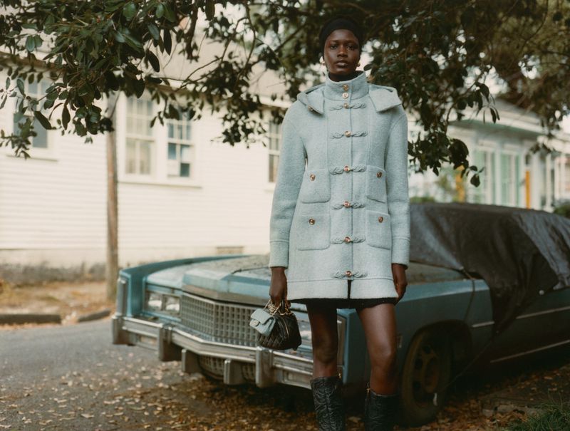 An adult woman poses in a hooded wool coat with a purse in an outdoor urban setting with a covered car and residential buildings.