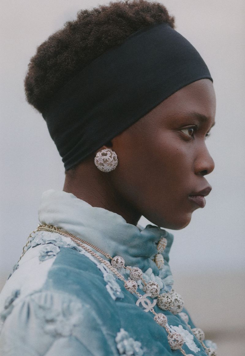 Close-up profile shot of a dark-skinned woman with a black headband, large round earrings, and an embellished Chanel jacket and necklace.