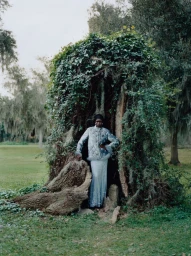 Model standing inside a large hollow tree covered in lush green vines wearing a patterned light-colored coat and skirt