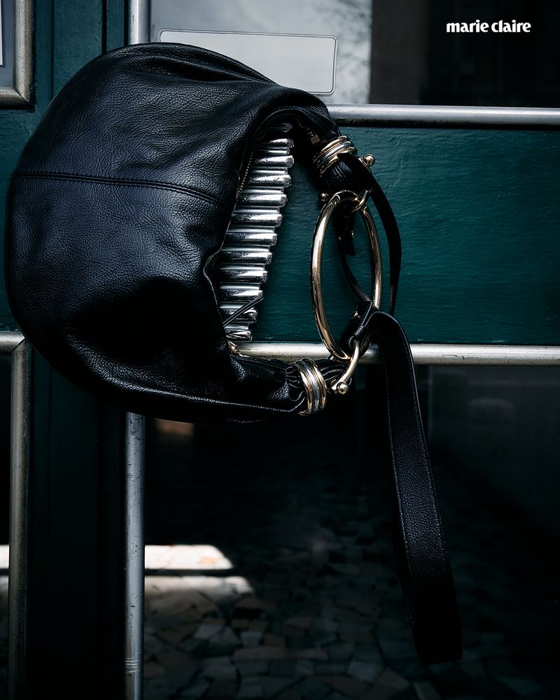 Black leather hobo bag with ring detail hanging on a silver metal railing against a dark teal door with the 'marie claire' logo in the upper right.