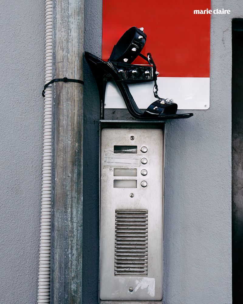 Black high-heel sandal with studs perched on an intercom system against a grey wall, illuminated by natural lighting and a magazine logo.