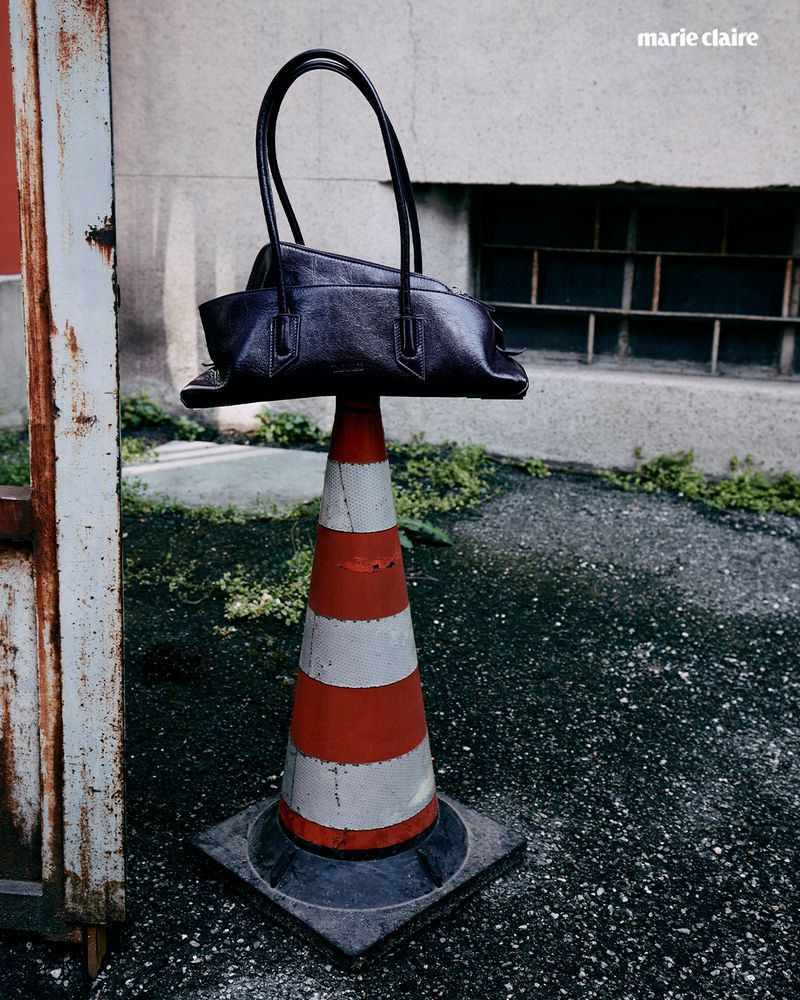 Fashion handbag on an orange-striped traffic cone outdoors near a weathered building and rusted metal door for a Marie Claire editorial.
