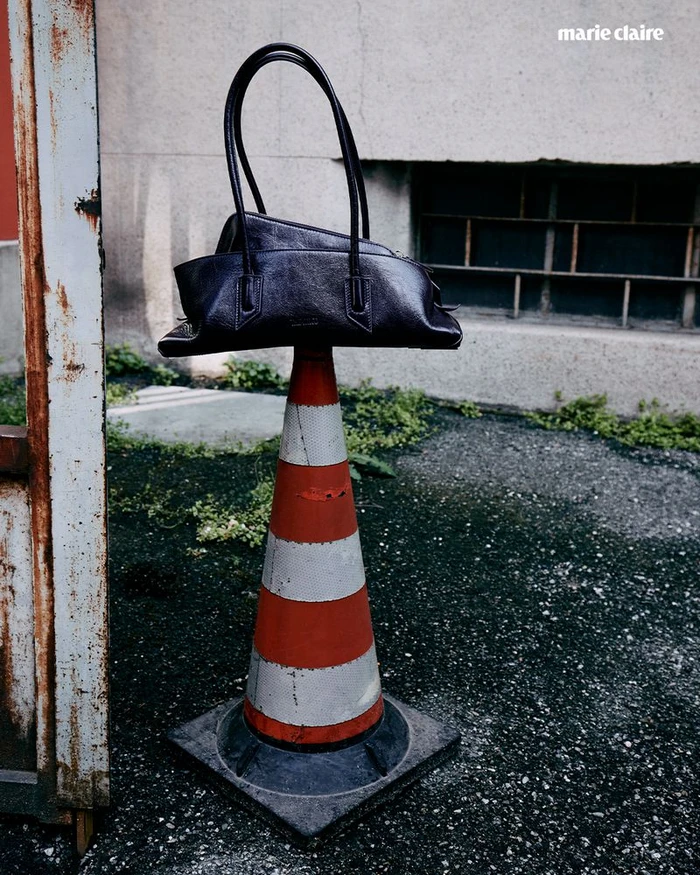 A black leather handbag balanced on top of an orange and white traffic cone outdoors against a concrete wall.