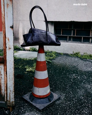 A black leather handbag balanced on top of an orange and white traffic cone outdoors against a concrete wall.
