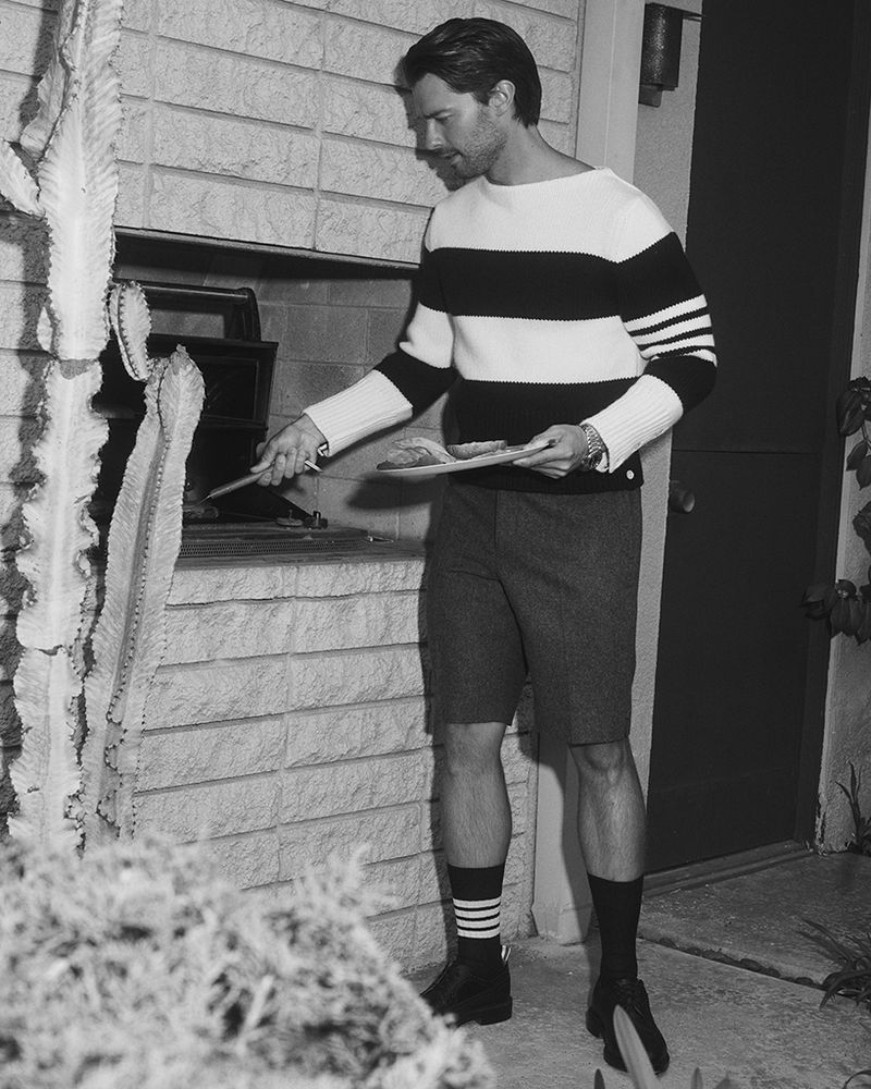 Monochrome image of a man in a striped sweater tending to a grill near a cactus, captured in an outdoor setting with natural light.