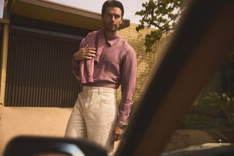 Portrait of a stylish adult man in a mauve shirt and cream pants, standing outdoors with a building and greenery behind.