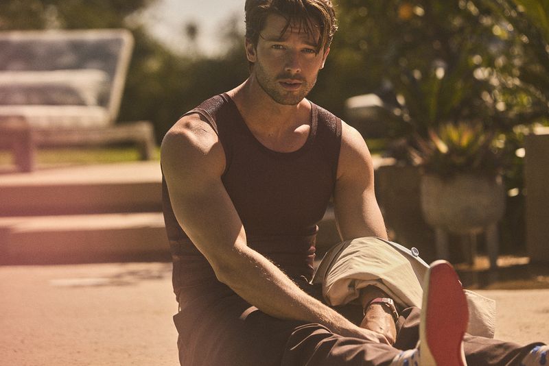 Attractive adult male in tank top sits outdoors in sunlight, looking towards the camera, medium shot, with blurred background of greenery.