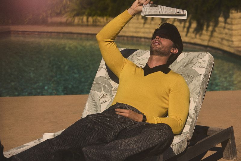 Man in yellow shirt lounges by pool with newspaper over eyes, resting in outdoor setting on a sunny day, looking relaxed and carefree.