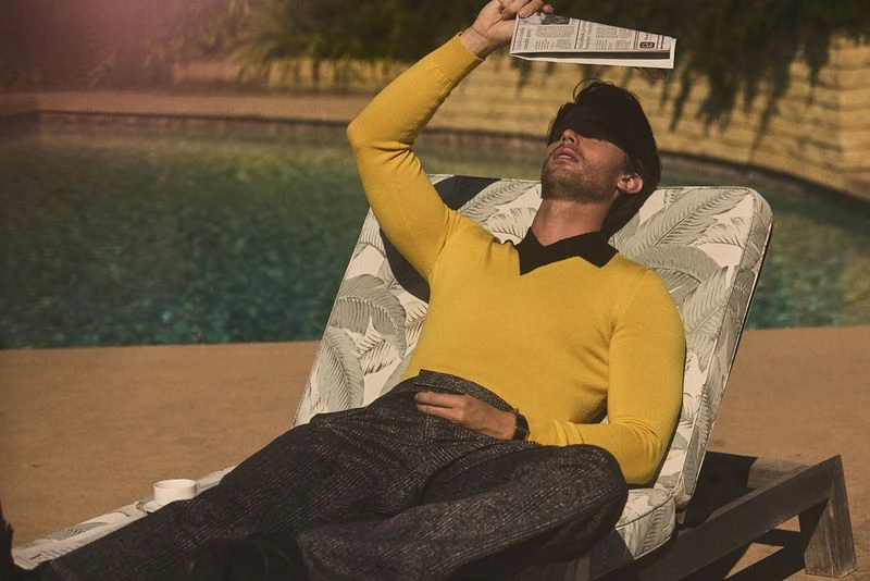Man in yellow shirt lounges by pool with newspaper over eyes, resting in outdoor setting on a sunny day, looking relaxed and carefree.