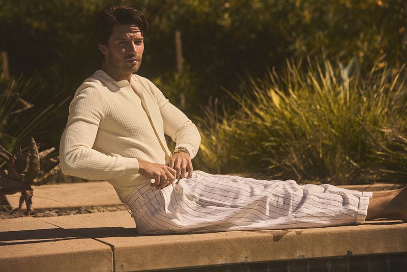 Adult male poses outdoors near a pool in a cream-colored shirt and striped pants, with greenery in the background and a neutral color palette.
