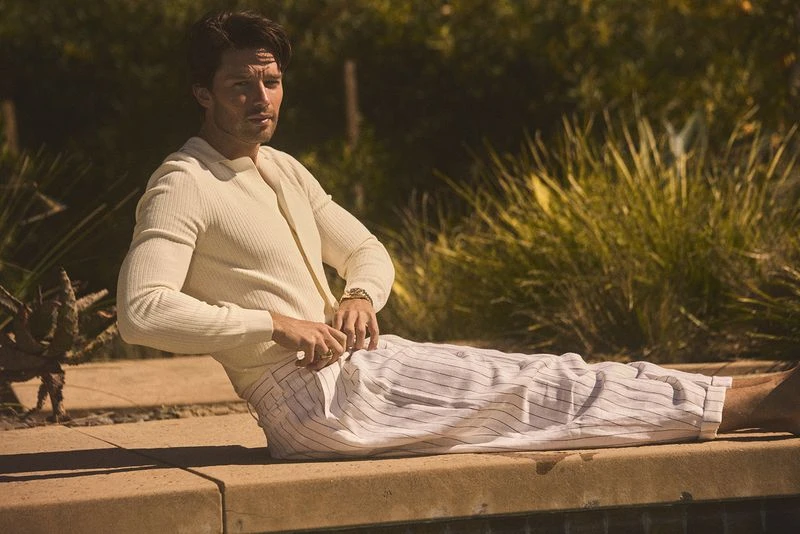 Adult male poses outdoors near a pool in a cream-colored shirt and striped pants, with greenery in the background and a neutral color palette.