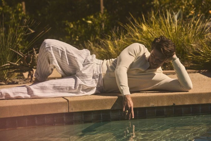 Man lies on side by pool in sunlight, wearing light-colored lounge pants and long-sleeve shirt, hand touches pool water, looking pensive.