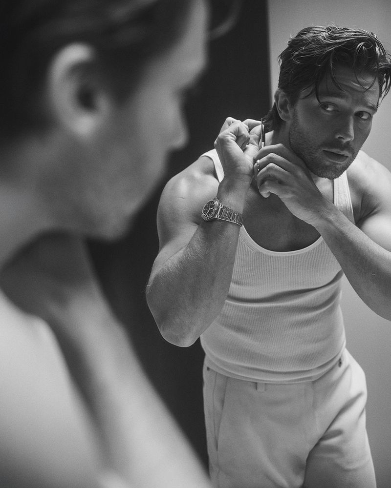 Black and white portrait of a Caucasian man in a tank top reflected in a mirror, shaving his neck with a straight razor.