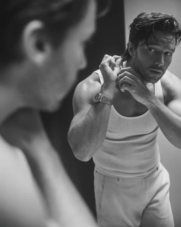 Black and white portrait of a Caucasian man in a tank top reflected in a mirror, shaving his neck with a straight razor.