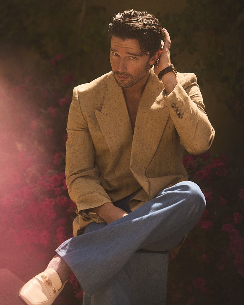 Portrait of handsome man in a tan blazer and blue jeans sitting outdoors among dark green foliage with bright pink flowers.
