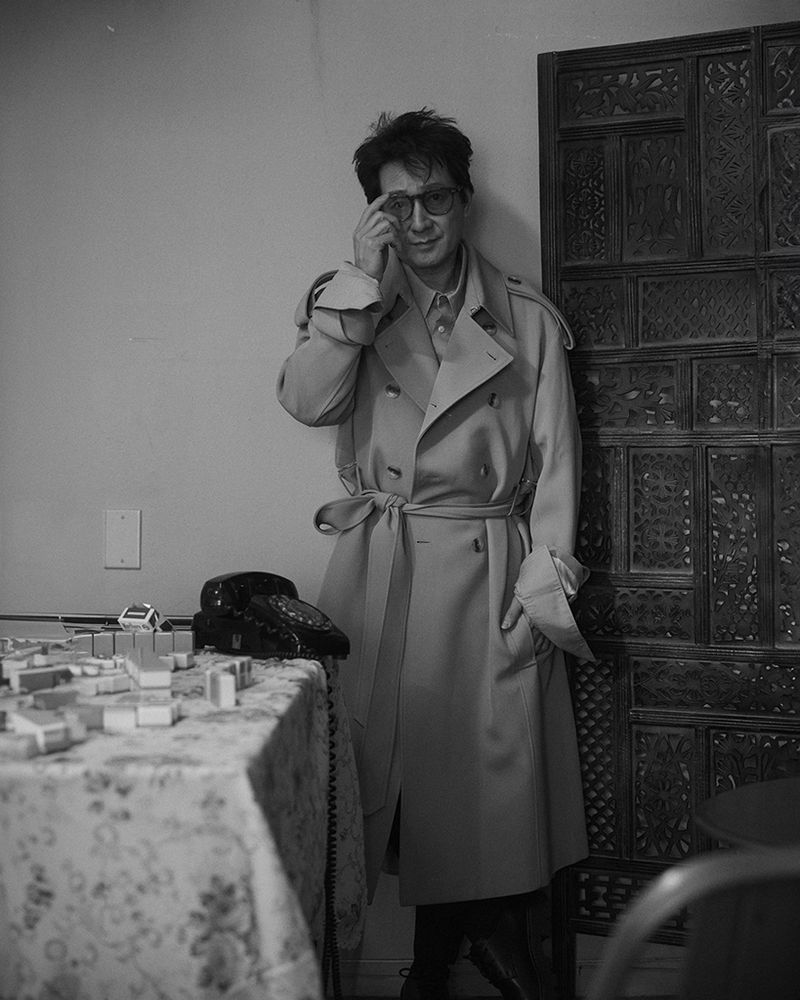 Monochrome portrait of a man with glasses and a trench coat standing next to a decorated partition with a vintage telephone and small objects on a table.