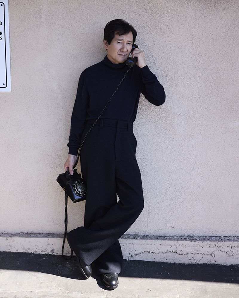 Adult male in black turtleneck and wide-leg pants leans against a wall holding a rotary phone and receiver to his ear indoors.