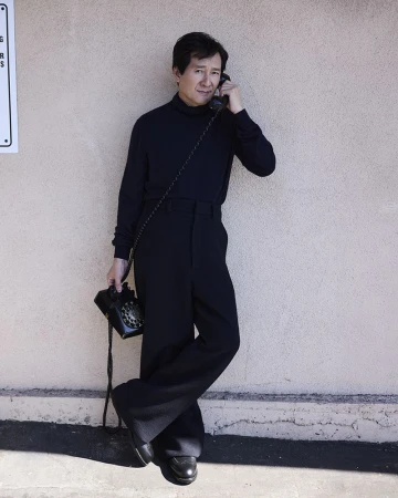 Adult male in black turtleneck and wide-leg pants leans against a wall holding a rotary phone and receiver to his ear indoors.
