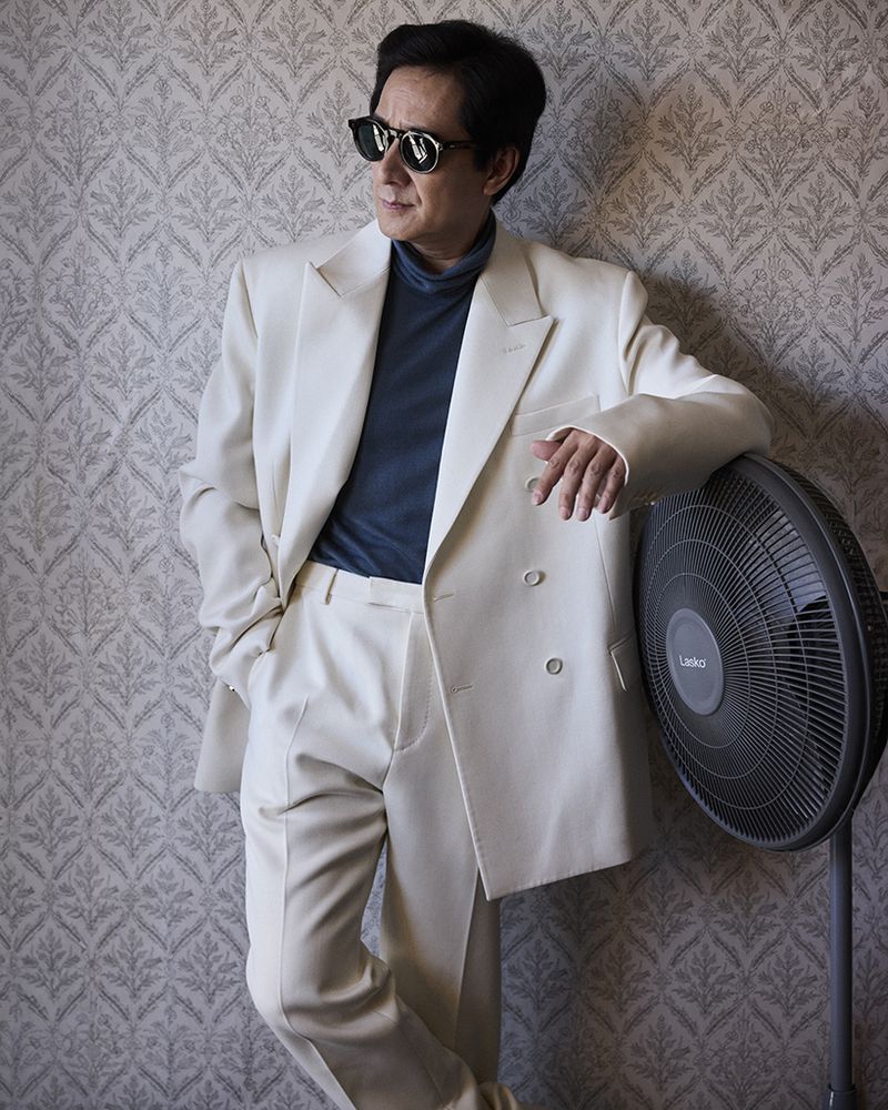 Elegant man in a white suit and sunglasses leans against a patterned wall with a fan; indoor setting with soft, diffused lighting.