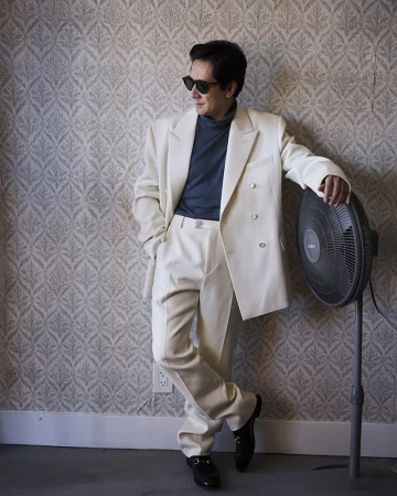 Adult male in a white suit, black shoes and sunglasses leans against wall with a fan in an indoor setting with patterned wallpaper.