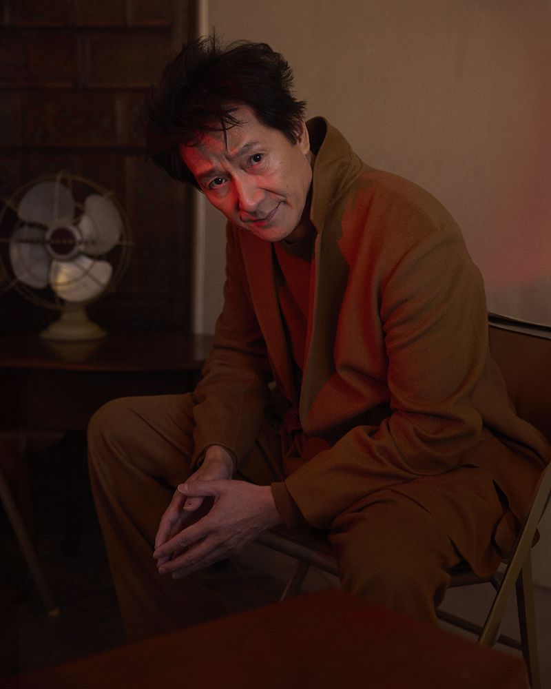 Portrait of an Asian man in his 60s with dark, messy hair, wearing a brown suit, sitting on a chair in a dimly lit indoor setting.