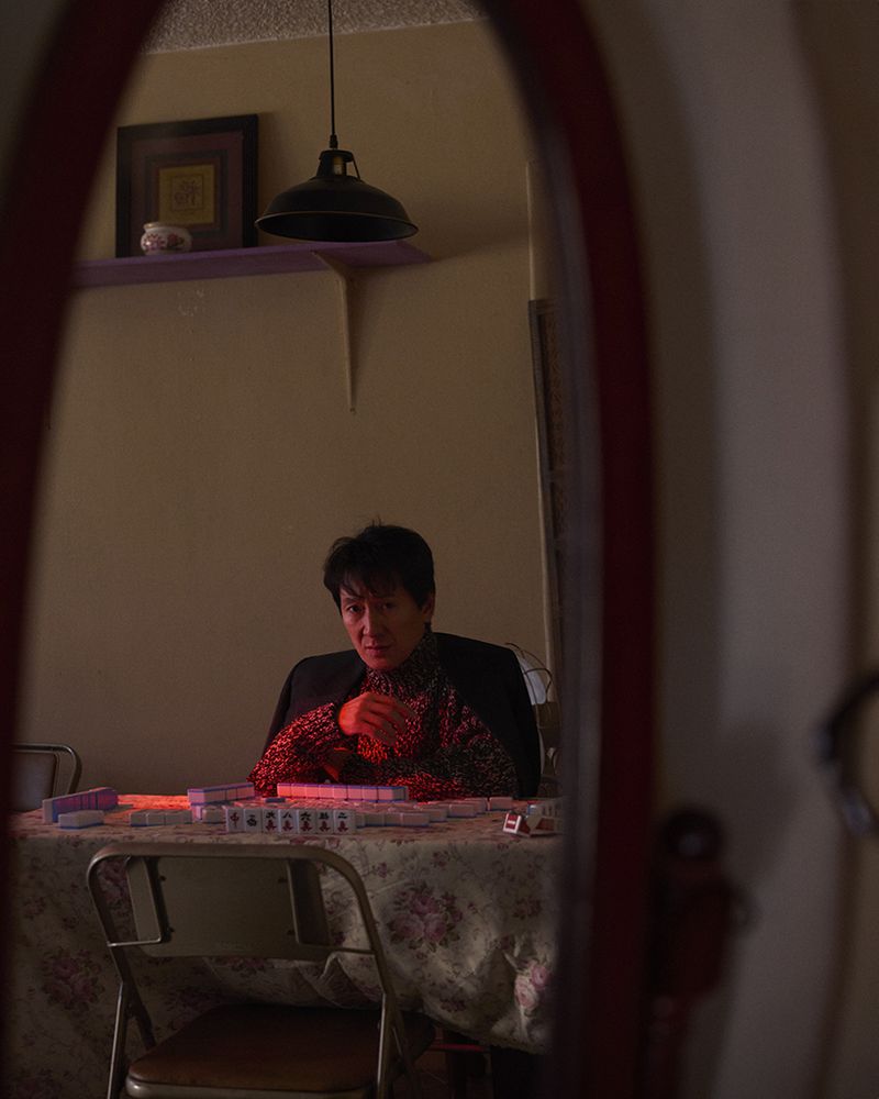 Person sitting at a table covered in mahjong tiles, illuminated by a red light, reflected in an oval mirror indoors.