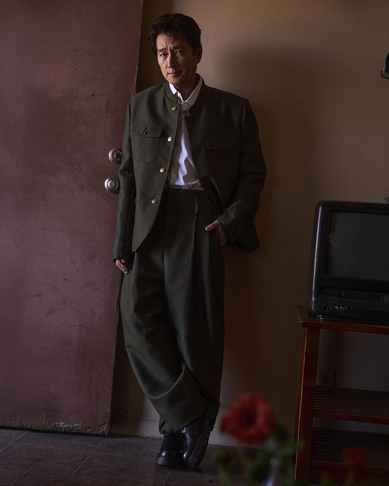 Adult man in dark green suit and black shoes leans against a door inside a home with TV and red flowers, looking at camera.