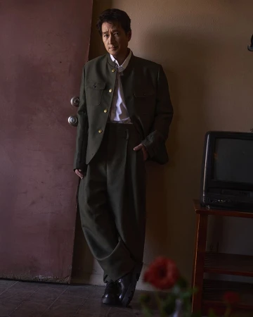 Adult man in dark green suit and black shoes leans against a door inside a home with TV and red flowers, looking at camera.