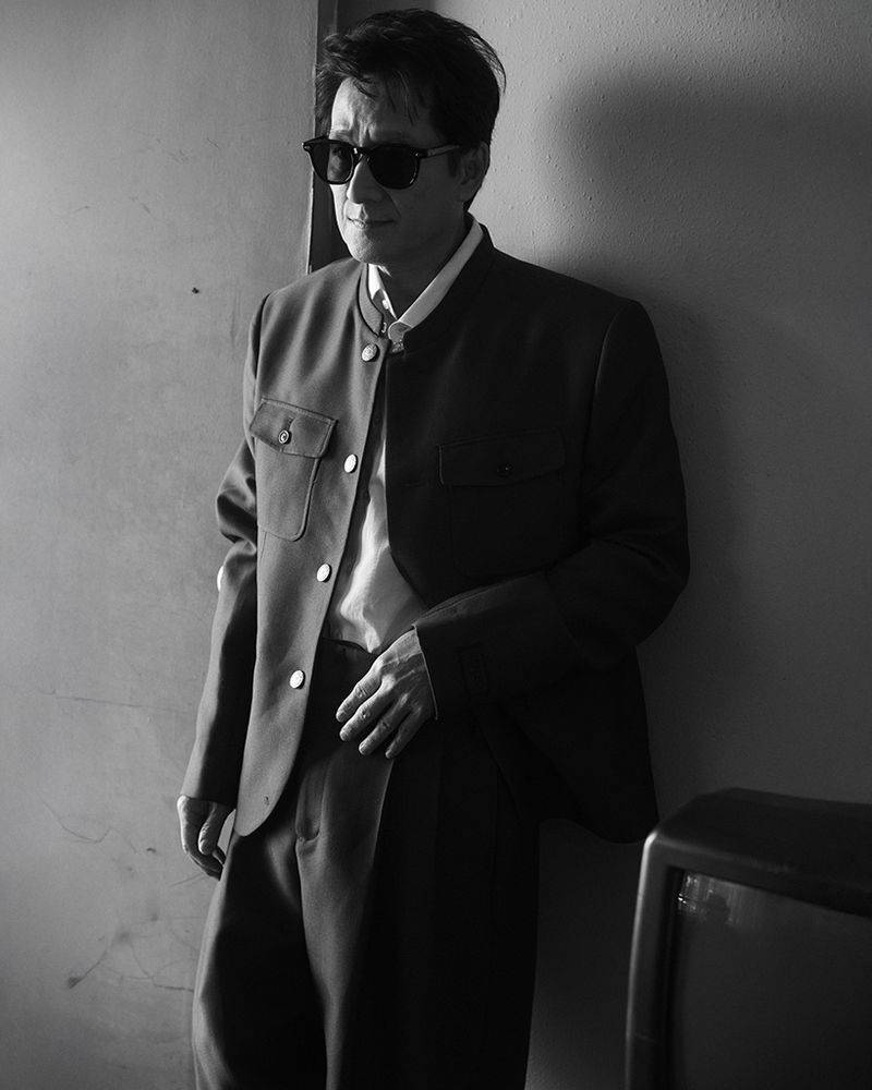 Black and white shot of an adult male wearing sunglasses, a jacket, and dress pants, standing in an indoor setting next to a wall.