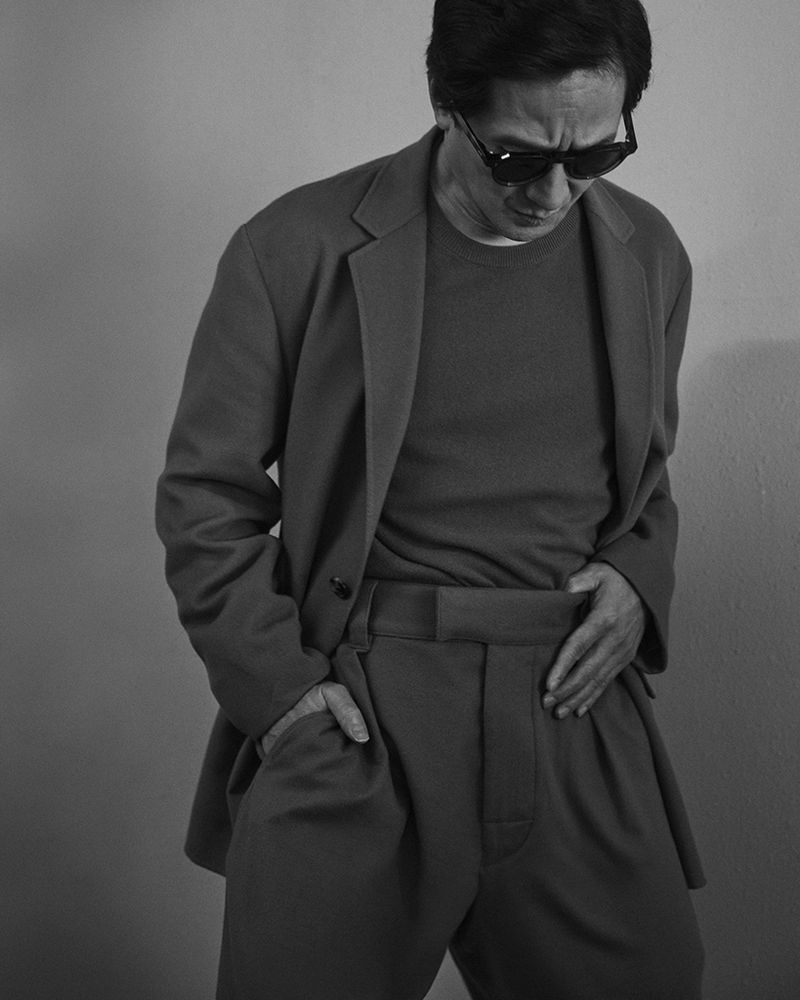Black and white portrait of a man in a suit and sunglasses with his hand in his pocket, gazing downward against a grey background.