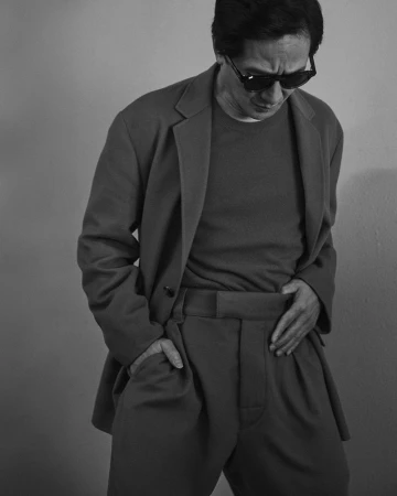 Black and white portrait of a man in a suit and sunglasses with his hand in his pocket, gazing downward against a grey background.