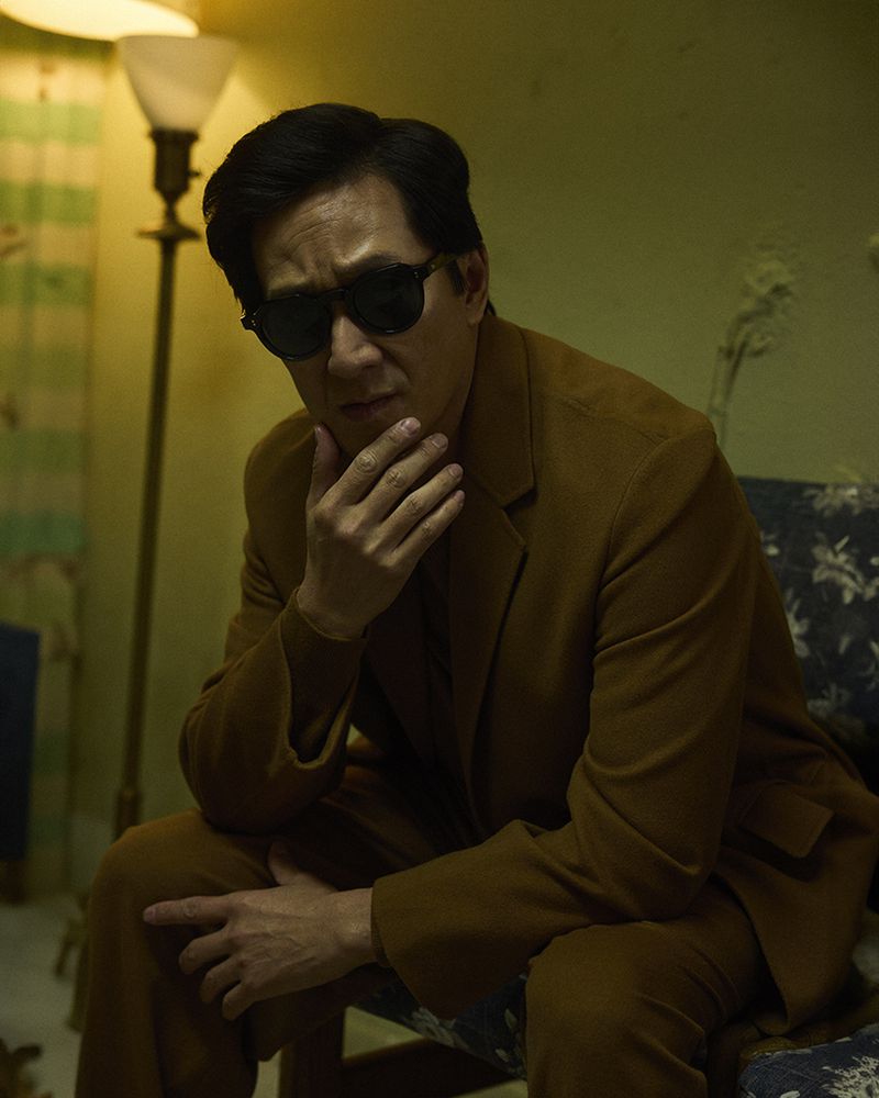 Portrait of an adult Asian man in a brown suit and dark sunglasses, seated indoors with a neutral facial expression and hand to his chin.