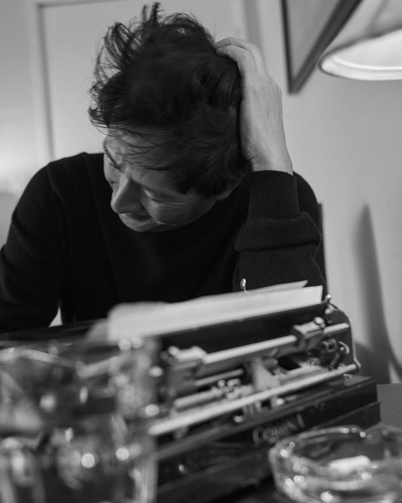 Black and white portrait of a seated adult man with disheveled hair, his hand on his head, next to a vintage typewriter.
