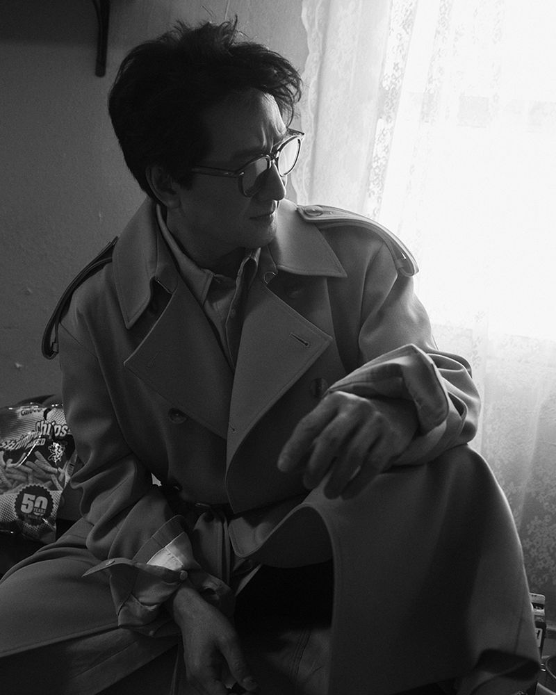 Monochrome portrait of a man wearing glasses and a trench coat sitting in front of a window with lace curtains, looking to the side.