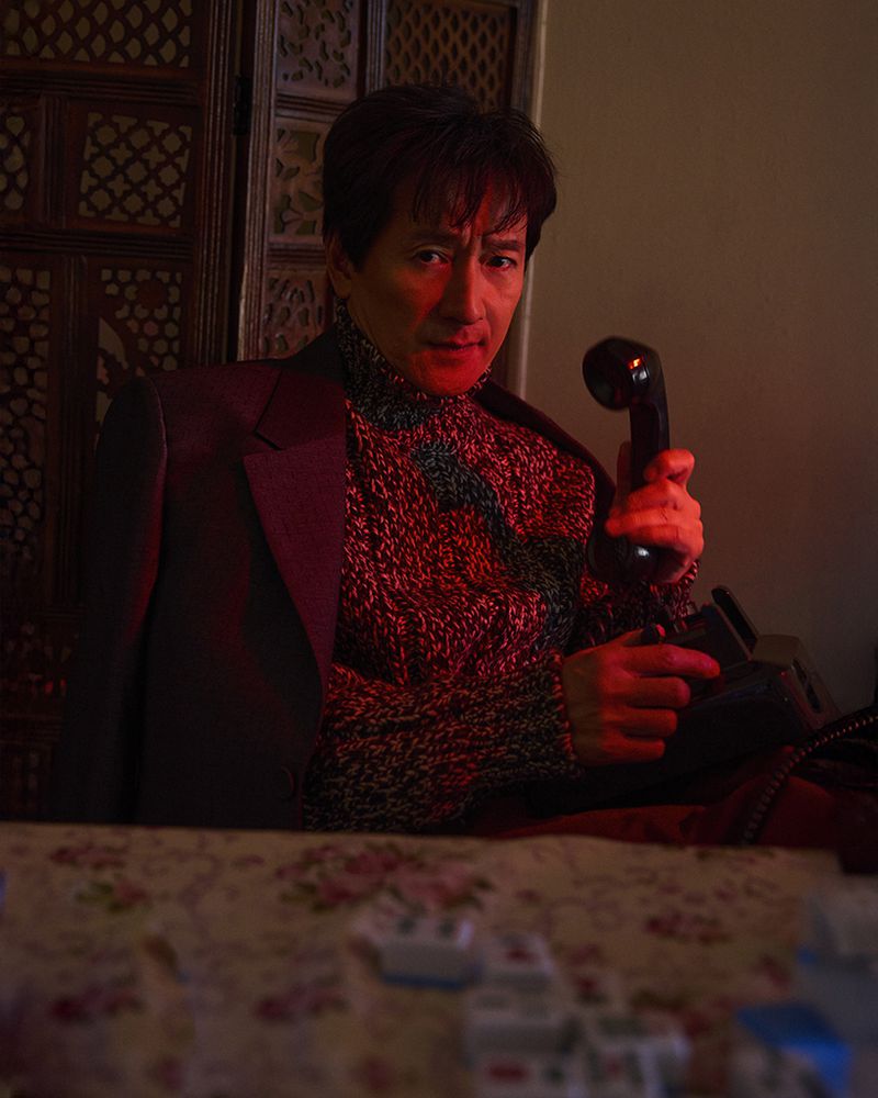 Adult male in sweater and suit jacket speaks into vintage telephone; indoor setting with floral table and ornately carved wall.