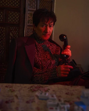 Adult male in sweater and suit jacket speaks into vintage telephone; indoor setting with floral table and ornately carved wall.