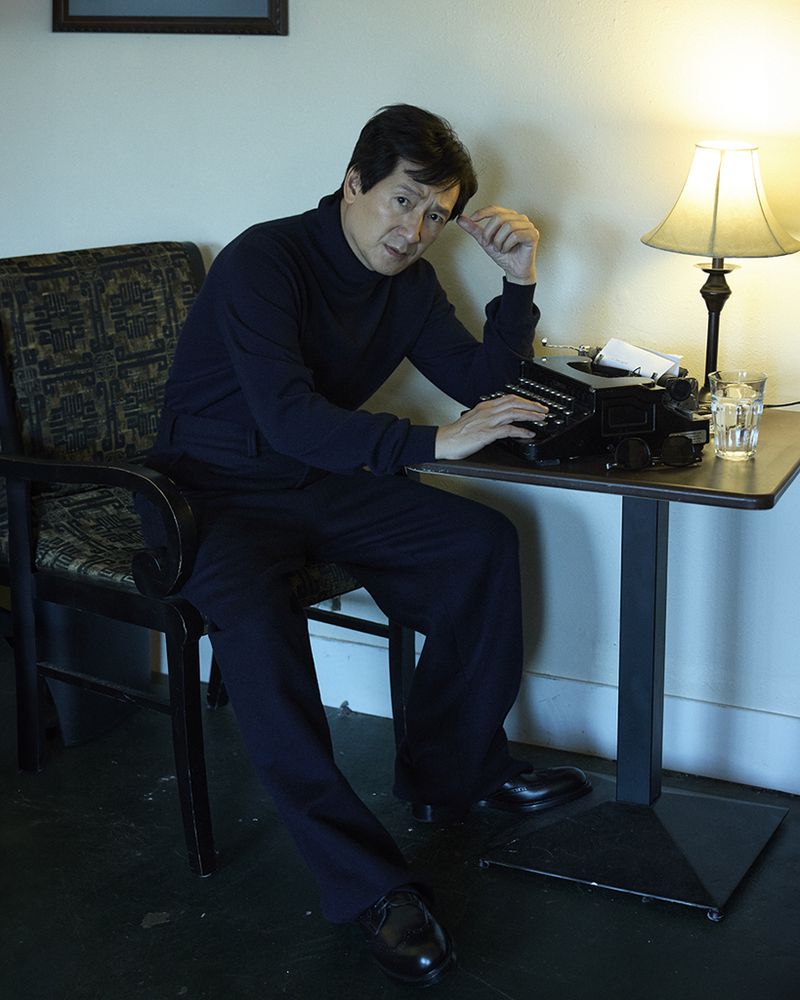 An Asian man is sitting in a chair next to a table, looking directly at the camera, with a typewriter and lamp on the table.
