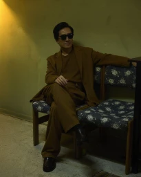 Ke Huy Quan seated on patterned chairs wearing a brown suit and sunglasses in a warmly lit room for Man About Town.