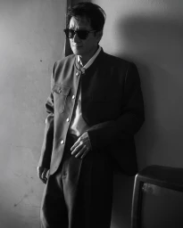 Ke Huy Quan wearing dark sunglasses and a buttoned jacket leaning against a plain wall in a black and white portrait.