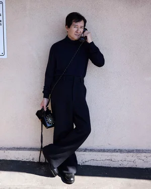 Ke Huy Quan wearing a black turtleneck and trousers holding a vintage rotary telephone against a textured wall