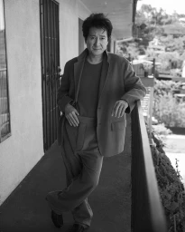 Ke Huy Quan leaning against a balcony railing wearing a tailored suit jacket and trousers in a monochrome editorial portrait