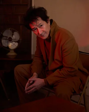 Ke Huy Quan seated on a chair wearing a brown suit jacket in a dimly lit room with a vintage fan