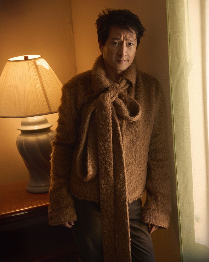 Ke Huy Quan wearing a brown textured jacket with a large scarf tie, posing next to a lit table lamp for Man About Town.