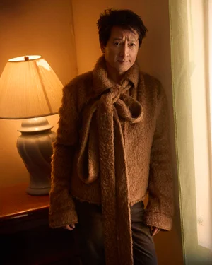 Ke Huy Quan wearing a brown textured jacket with a large scarf tie, posing next to a lit table lamp for Man About Town.